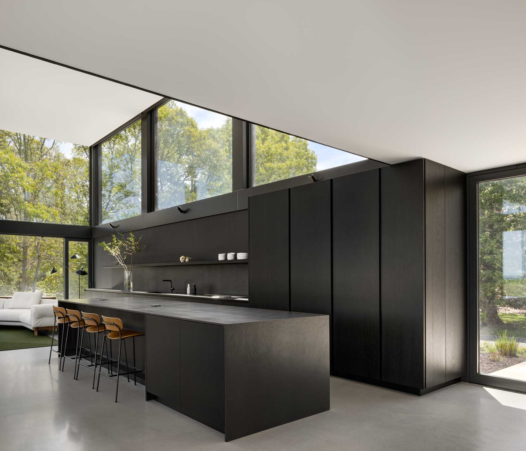 Above this modern black kitchen, large glazed openings act as a clerestory and flood the space with diffuse natural light, framing views of the surrounding treetops.